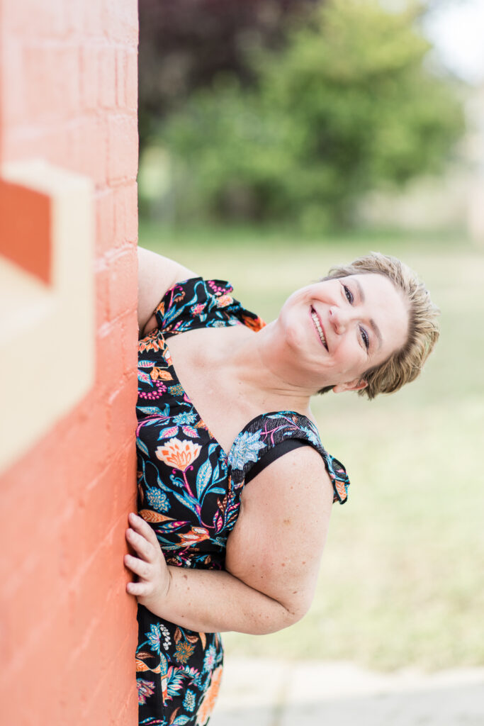 Create your Space with Shara - Shara in a colourful jumpsuit peering around the corner of a building and smiling at the camera.