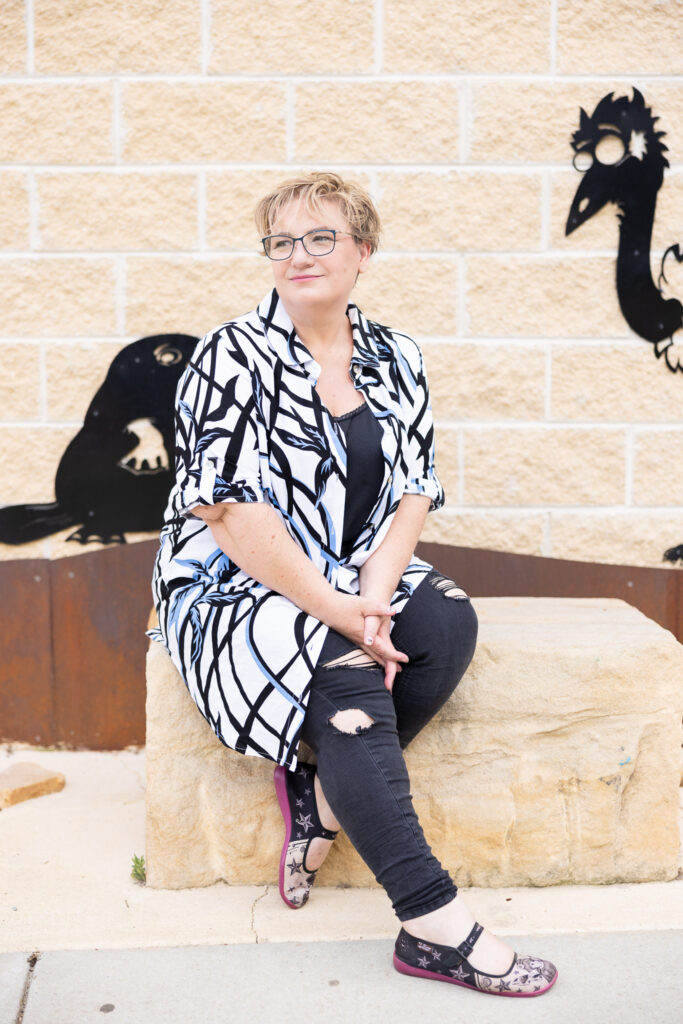 Create your Space with Shara - Shara wearing a patterned shirt and glasses, sitting on a sandstone block, looking to the left.