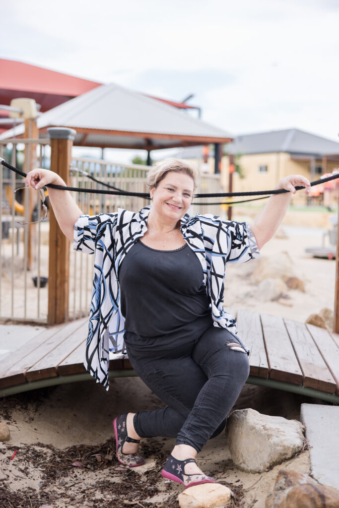 Create your Space with Shara - Shara wearing a colourful shirt sitting on a small bridge in a children's playground with arms stretched along the post rope.