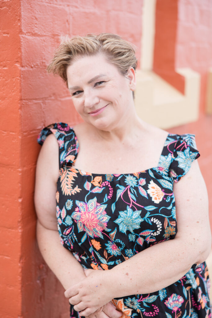 Shara wearing a colourful jumpsuit, leaning against the edge of an orange painted brick wall.