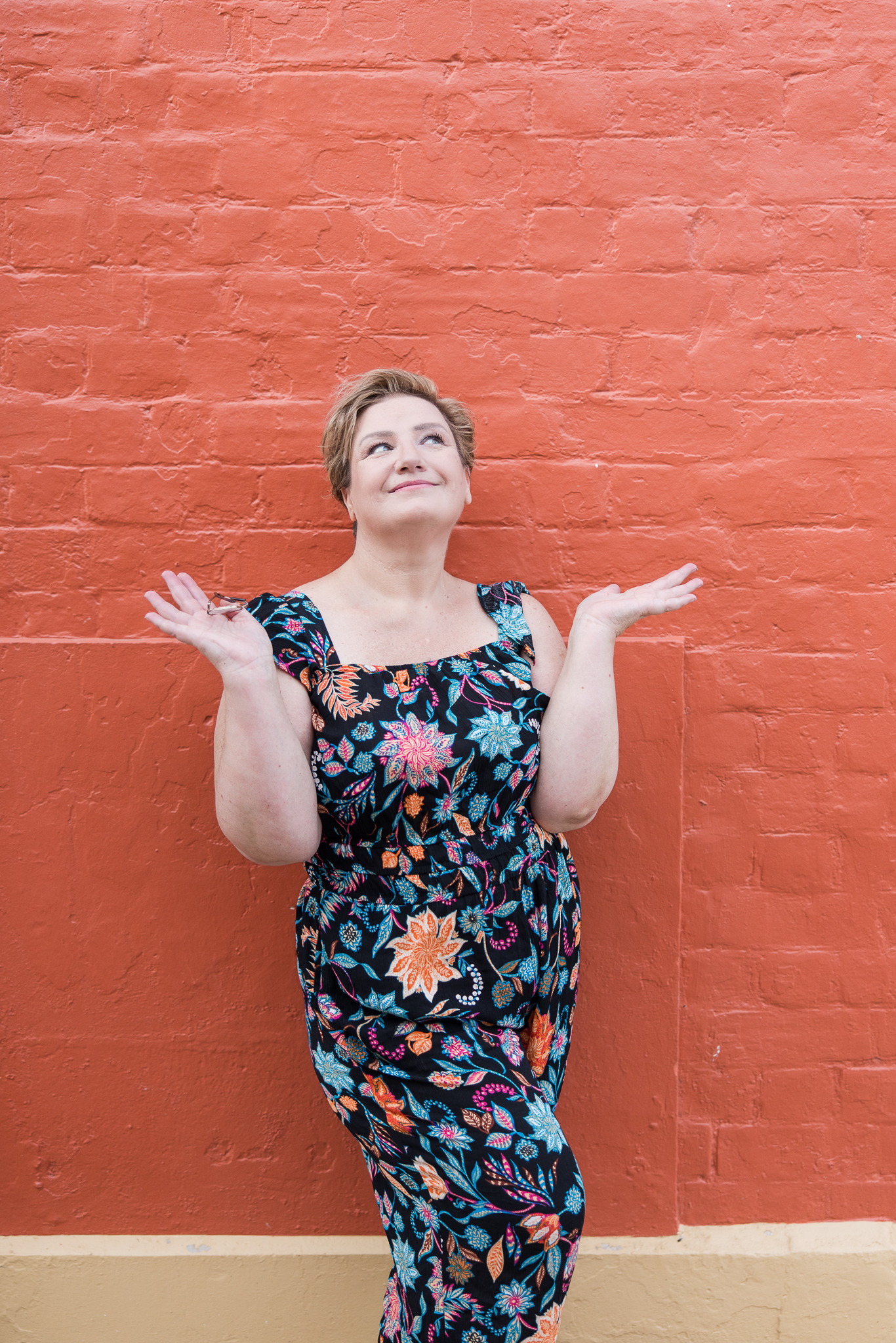 Create Your Space With Shara - Shara wearing a colourful jumpsuit next to an orange painted brick wall, raising her hands.