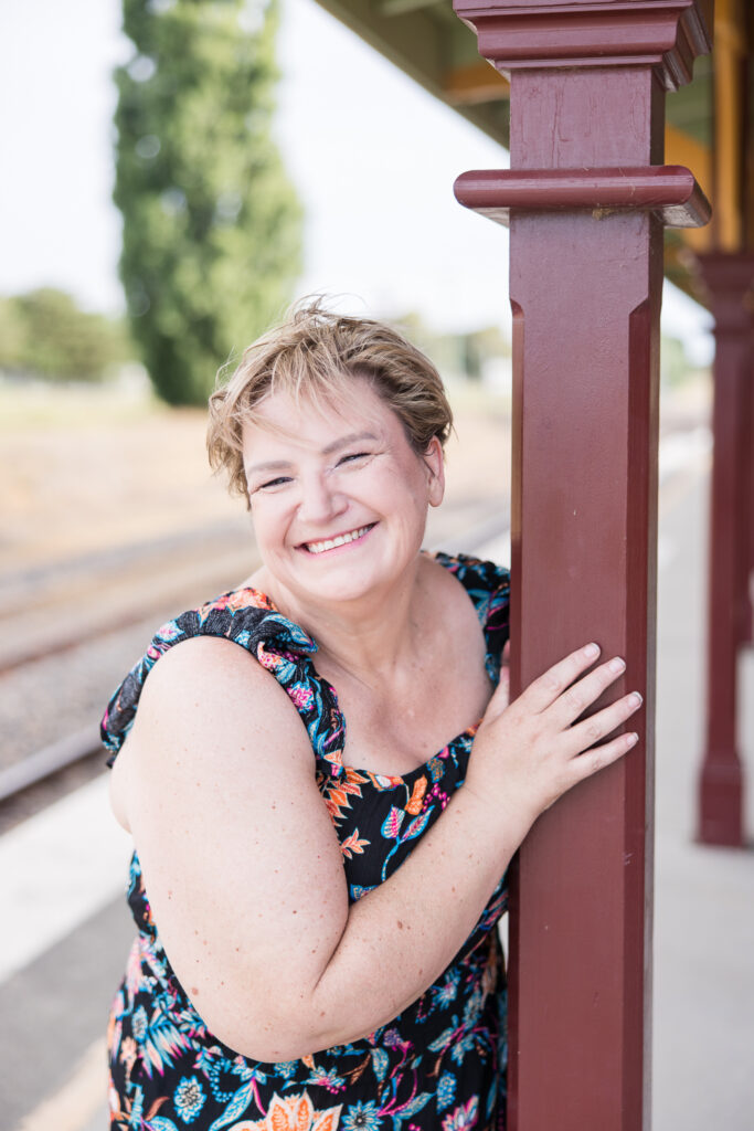 Create your Space with Shara - Shara  wearing a colourful jumpsuit, leaning against a pole at a train station and smiling at the camera.