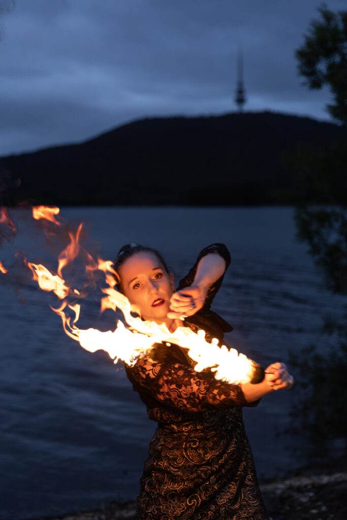 Photograph of a woman standing next to a lake and fire twirling 